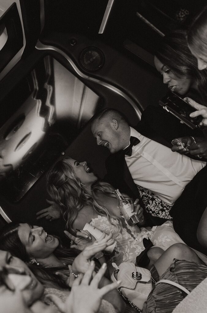A black and white image shows a bride and groom smiling and laughing in the back of a party bus on the way to their elopement at lakeside wedding and events